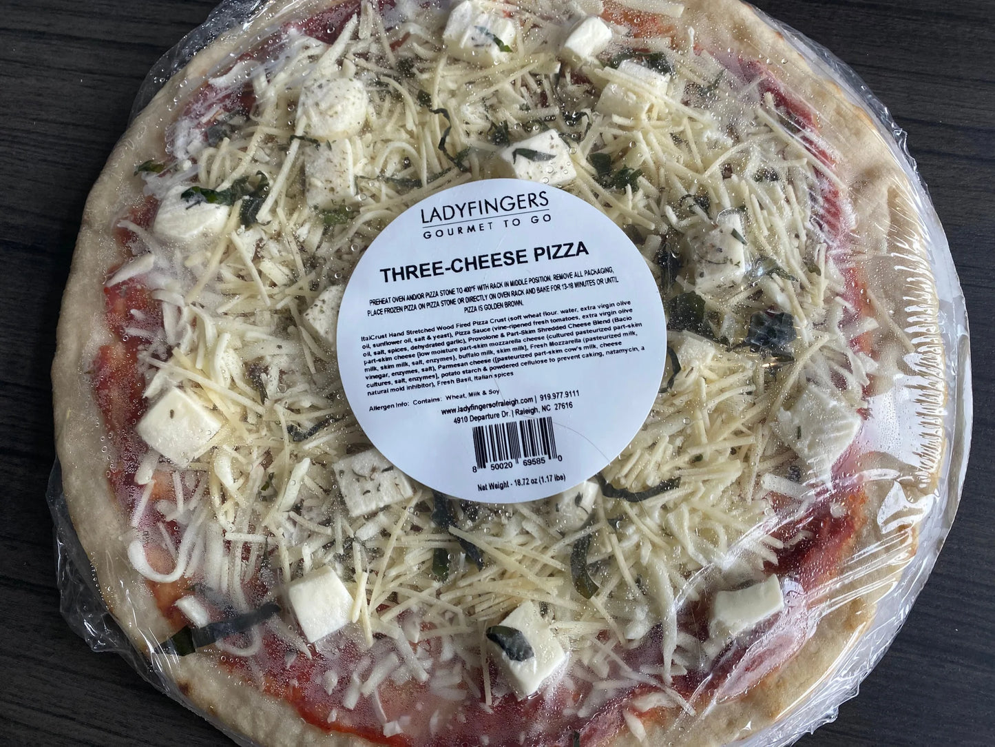 Three-cheese pizza with a label on top, wrapped in plastic on a dark surface.