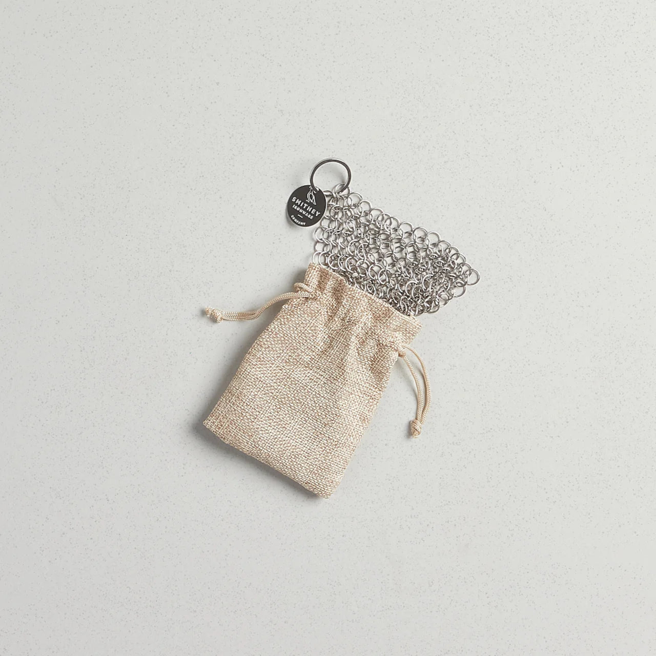 A chainmail scrubber is displayed on a beige burlap drawstring bag, indicating its use as a cleaning tool for cookware.