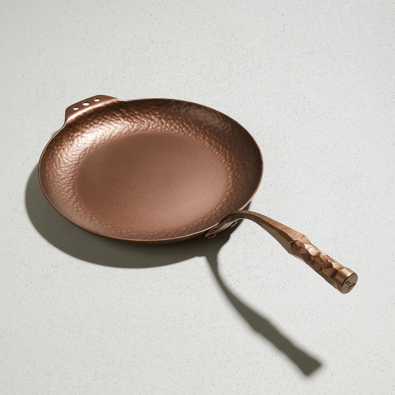 A bronze-colored carbon steel skillet with a hammer texture and a handle inspired by traditional fire tools, displayed on a light background.
