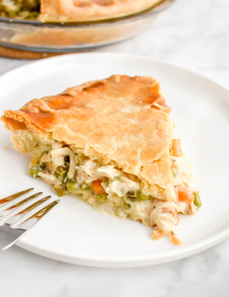 A slice of chicken and vegetable pie on a white plate with a fork, with the pie dish blurred in the background on a light marble surface.