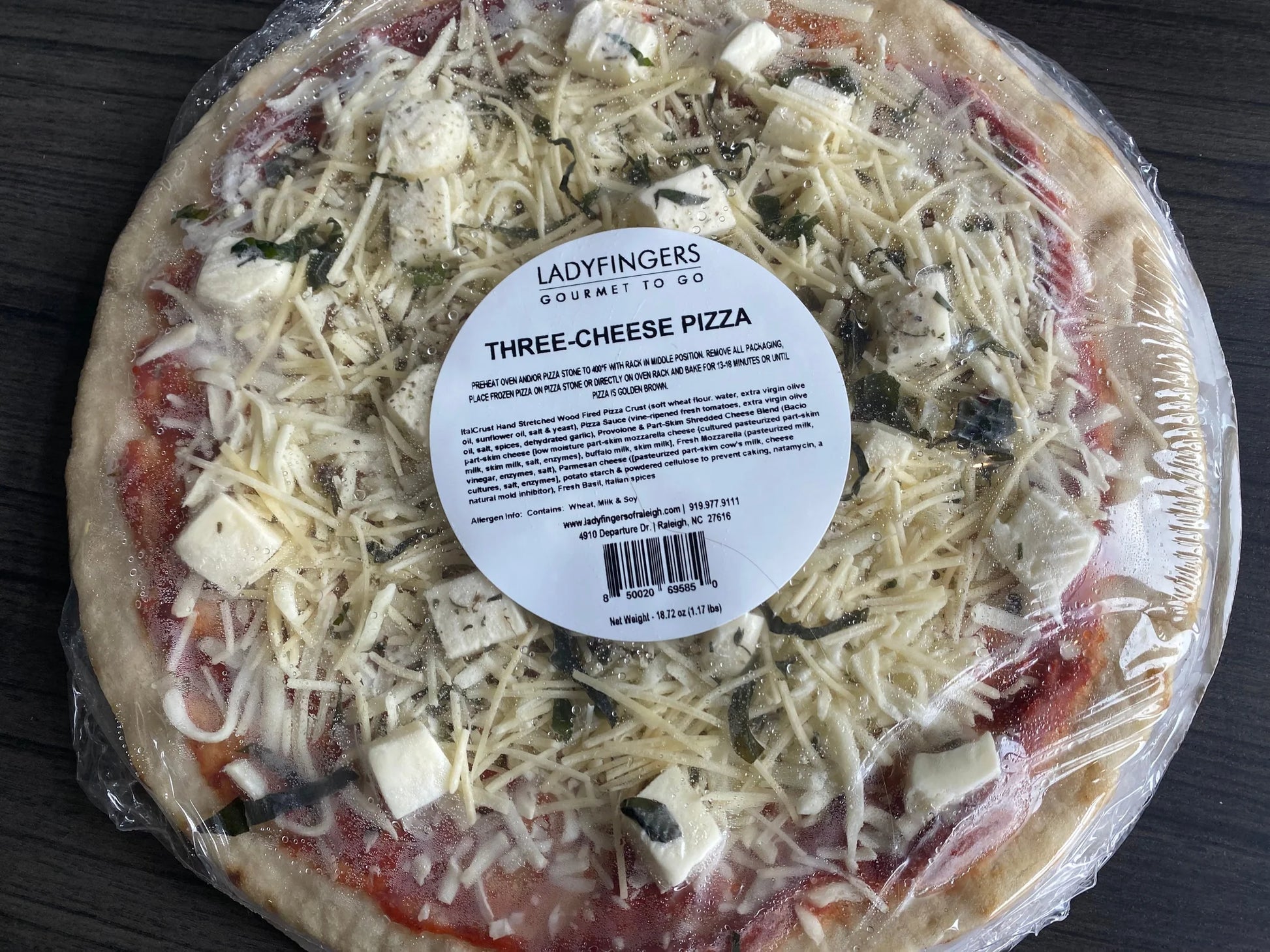 Three-cheese pizza with a label on top, wrapped in plastic on a dark surface.