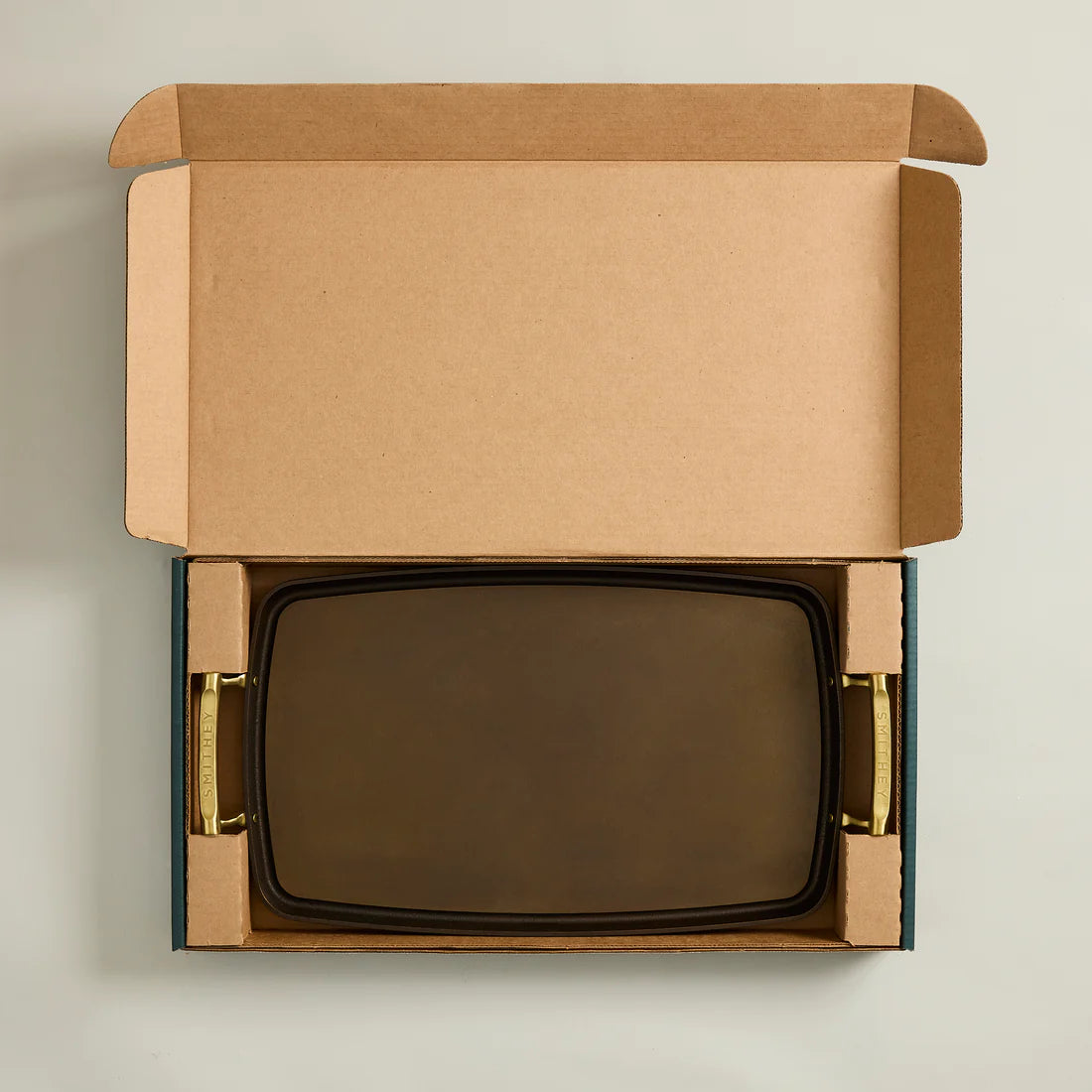 A polished, satin-smooth, brown metal griddle with embossed Smithey handles, packaged in a cardboard box.