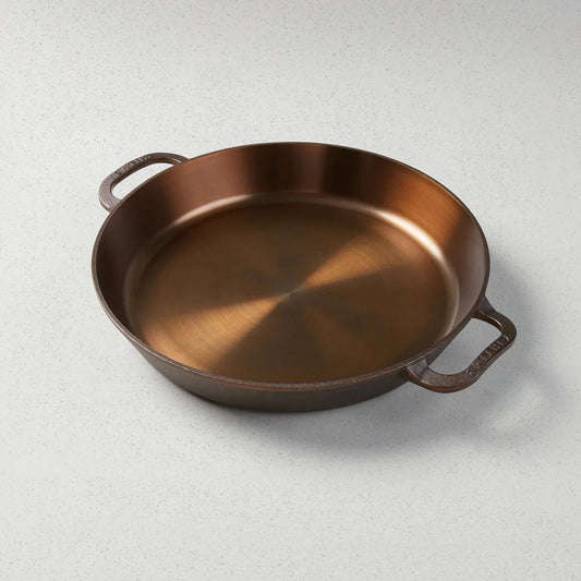 A bronze-colored cast iron skillet with two handles, viewed from an angle that highlights the skillet's generous size and depth.
