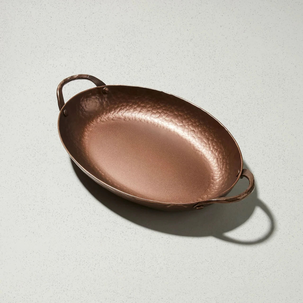 A hand-forged copper oval roaster with hammered handles, displayed on a light background.